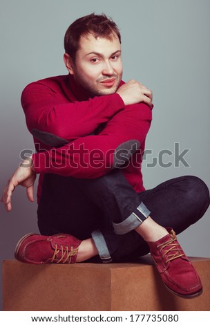 Male fashion concept. Portrait of a young man wearing trendy casual purple sweater, blue jeans and purple shoes. Guy sitting on a wooden cube & posing over gray background. Hipster style. Studio shot
