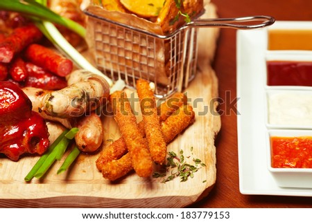 Fried cheese sticks with small meat sausages served with scallion and dried herbs on wooden cutting board with a set of marinara, mayonnaise, honey-mustard sauces and ketchup.