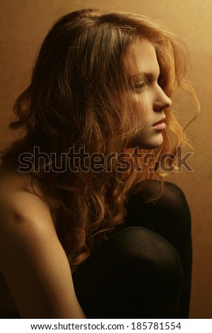 Emotive portrait of a young beautiful girl with red hair in profile hugging her knees and posing over wooden background. Close up. Studio shot