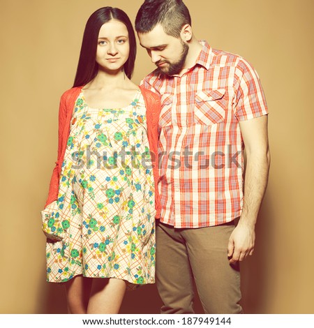 Stylish pregnancy concept: portrait of two happy hipsters (husband and wife) in trendy clothes (shirt, dress, cardigan and jeans) expecting their child. Pastel colors. Studio shot