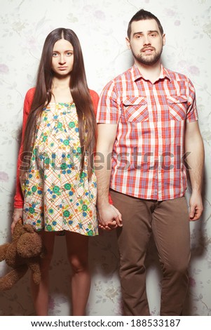 Stylish pregnancy concept: portrait of couple of hipsters (husband and wife) in trendy clothes (shirt, dress, cardigan and jeans) holding brown teddy bear. Vintage (retro) style. Studio shot