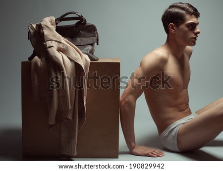 Male high fashion concept. Portrait of a handsome male model sitting near a wooden cube in trendy underwear. Cardigan and leather bag on a cube. Perfect skin & haircut. Vogue style. Studio shot