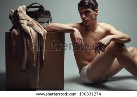 Male high fashion concept. Portrait of a handsome male model sitting near a wooden cube in trendy underwear. Cardigan and leather bag on a cube. Perfect skin & haircut. Vogue style. Studio shot