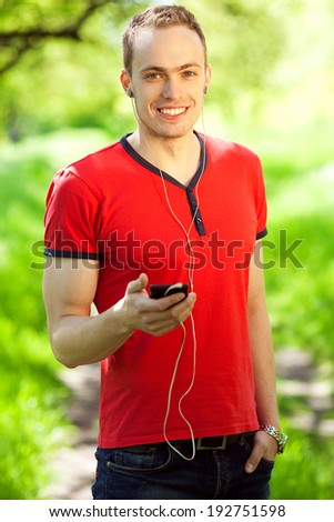Gadget freak concept. Portrait of a young muscular man in red t-shirt and jeans walking with cellphone in the park. White shiny smile. Sunny weather. Outdoor shot