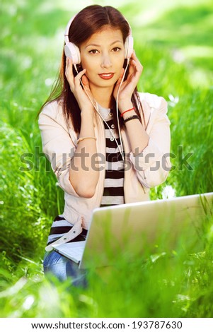 Happy fashionable hipster girl in pink leather jacket listening to the music on computer, sitting in green grass in the park. Sunny day. Outdoor shot