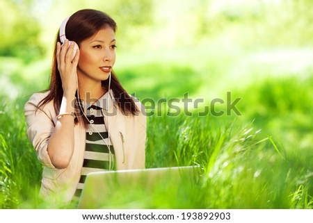 Happy fashionable hipster girl in pink leather jacket listening to the music on computer, sitting in green grass in the park. Sunny day. Copy-space. Outdoor shot