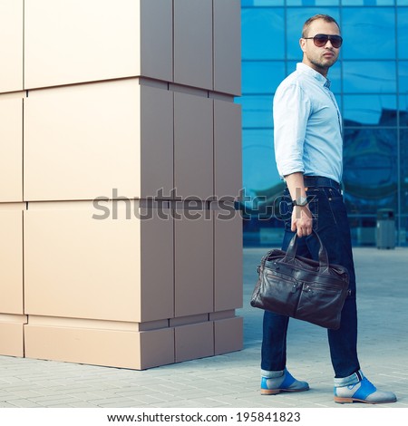 Metrosexual Man Concept. Portrait of attractive man in trendy casual clothing with leather bag and sunglasses posing over shopping mall. Sunny spring weather with blue sky. Outdoor shot
