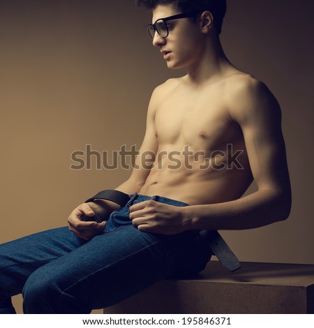 Blue Jeans & Eyewear Concept. Male beauty. Handsome male model with nice abs in jeans and trendy glasses. Boy sitting on a wooden cube. Hipster style. Fashion studio portrait