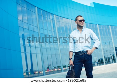 Men Shopping concept. Portrait of attractive smiling man in trendy casual clothing with leather bag and sunglasses posing over shopping mall. Sunny spring weather with blue sky. Outdoor shot
