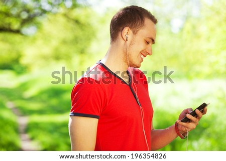 Profile portrait of a smiling young man in red t-shirt using smartphone in the park. Handsome muscular guy in casual clothing. Sunny summer day. Close up. Copy-space. Outdoor shot