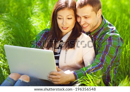 Happy weekend concept. Portrait of a hipster couple in trendy casual clothing sitting in green grass in park, using a laptop and looking happy together. Sunny spring (autumn) weather