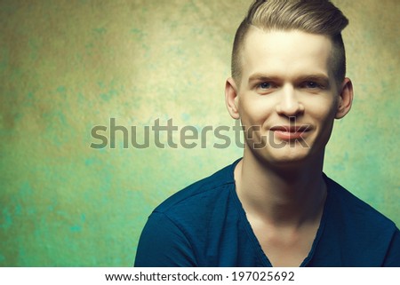 Hipster haircut concept. Portrait of young man in blue t-shirt with stylish haircut posing over golden-turquoise  background and smiling. Perfect skin & hair. Close up. Copy-space. Studio shot