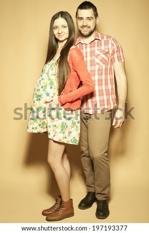Stylish pregnancy concept: portrait of two happy hipsters (husband and wife) expecting their child posing in trendy clothes (shirt, dress, cardigan, jeans and leather boots). Studio shot