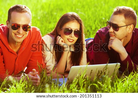 Useful weekend concept. Three happy friends having fun in the autumn park, lying together in green grass and using laptop. Trendy casual clothing and stylish eyewear. Shiny weather. Outdoor shot