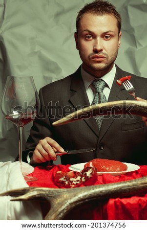 Baroque supper of blue blood aristocrat. Portrait of handsome man eating fresh meat at the vintage restaurant. Baroque accessories: deer horn, red and white table sheet. Indoor shot