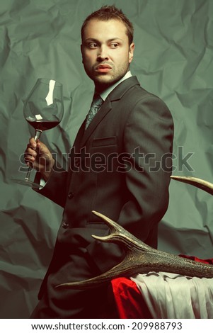 Emotive portrait of handsome stylish man in elegant dark gray suit drinking red wine. Baroque accessories: deer horn, red and white cloths. Retro style. Studio shot