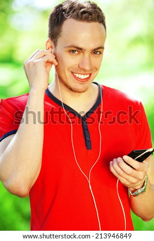 Portrait of happy young man in red t-shirt listening to music on smartphone in park. Handsome muscular guy in casual clothing. Sunny summer day. Street casual style. White shiny smile. Outdoor shot