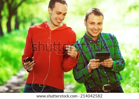 Gadget user concept. Funny hipster guys in trendy casual clothing reading e-book, listening music in park. White shiny smile. Bristle on face with healthy skin. Hipster style. Outdoor shot