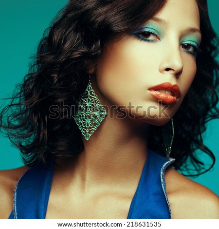 Disco party girl concept. Portrait of fashionable model with curly hair and arty make-up posing over blue background. Vintage earrings. Perfect skin. Retro style. Close up. Studio shot