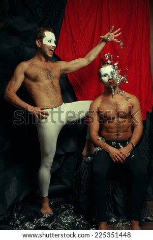 Hocus pocus concept. Two muscular mime artists, clowns with white masks on faces performing focus sequins and red apple over red cloth & black background. Magician and victim of trick. Studio shot