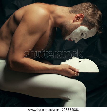 Mystery double concept. Arty portrait of circus performer in tights holding and looking at venetian mask (volto bianco), posing over black cloth. Muscular body and perfect tan. Studio shot