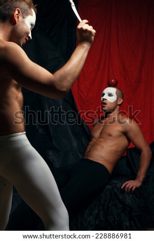 Bulls eye concept. Two muscular mime artists, clowns with white masks on faces performing trick with knife and apple over red cloth & black background. Studio shot