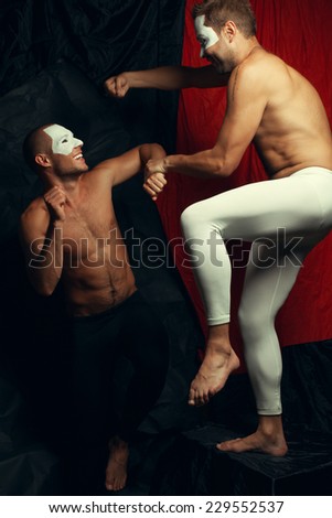 Freak circus concept. Two muscular mime artists, clowns with white masks on faces fighting and laughing over red cloth & black background. Vintage style. Studio shot