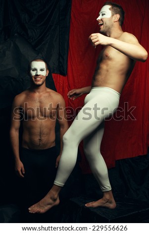Freak circus concept. Two muscular mime artists, clowns with white masks on faces playing fools and posing over red cloth & black background. Vintage style. Studio shot
