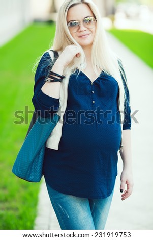 Stylish Pregnancy Concept. Portrait of fashionable mommy with long blond hair wearing casual trendy clothes, eyewear and going shopping with blue leather bag. Sunny weather. Outdoor shot