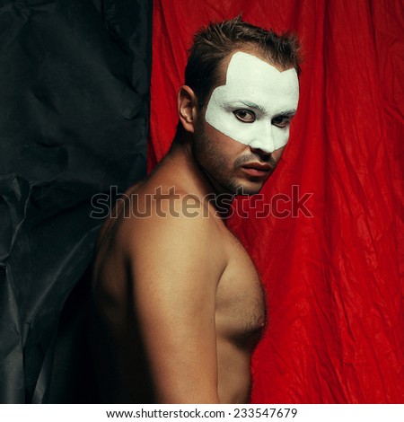 Backstage concept. Arty portrait of shirtless circus performer posing over black and red cloth. White mask on face. Muscular body and perfect tan. Halloween party. Studio shot