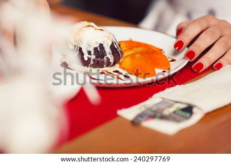 Haute cuisine concept. Woman\'s hand with perfect manicure and ice cream brownie sundae with chocolate sauce and slices of date plum on white plate. Close up. Indoor shot