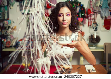 Winter holidays celebration concept. Portrait of doll like brunette in retro polka-dot sleeveless dress drinking tea or coffee in restaurant, cafe. Homemade christmas decoration. Close up. Indoor shot
