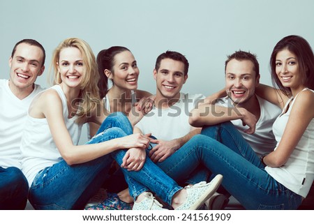 Happy together concept. Group portrait of healthy boys and girls in white t-shirts, sleeveless shirts and blue jeans sitting and posing over gray background. Copy-space. Urban style. Studio shot