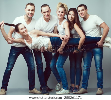 Happy together concept. Group portrait of healthy boys and girls in white t-shirts, sleeveless shirts and blue jeans holding friend in hands and posing over gray background. Urban style. Studio shot