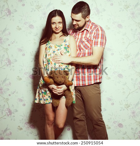 Stylish pregnancy concept: portrait of couple of hipsters (husband and wife) in trendy clothes (shirt, dress, jeans) holding teddy bear and smiling. Vintage (retro) style. Studio shot