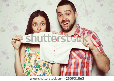 Stylish pregnancy concept: portrait of funny couple of hipsters (husband and wife) in trendy clothes (shirt, dress, jeans) holding small size baby shirt. Vintage (retro) style. Studio shot