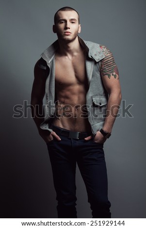 Portrait of tattooed brutal young man with short hair and bristle on face wearing sleeveless jacket, blue jeans and posing over gray background. Hipster style. Studio shot