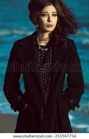 Emotive portrait of beautiful brunette with long curly hair in black coat. Luxurious golden accessories: earrings, necklace. Perfect make-up. Street & vogue style. Windy weather. Outdoor shot