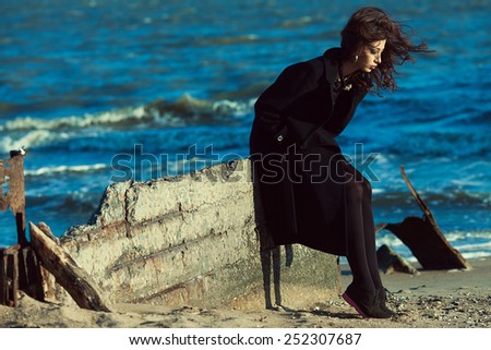 Full length portrait of beautiful brunette with long curly hair in black coat sitting at the seaside. Luxurious golden accessories: earrings, necklace. Street vogue style. Windy weather. Outdoor shot