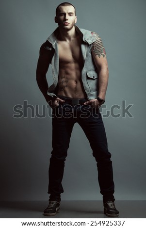 Full length portrait of tattooed brutal young man with short hair, bristle on face wearing sleeveless jacket, blue jeans, sneakers, posing over gray background. Hipster style. Studio shot