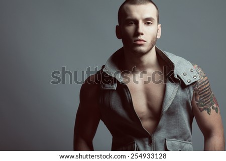 Portrait of tattooed brutal young man with short hair and bristle on face wearing sleeveless jacket and posing over gray background. Hipster style. Close up. Copy-space. Studio shot