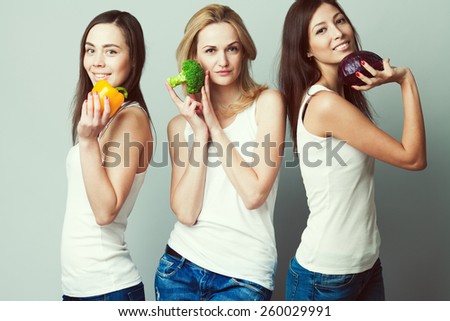 Happy veggies concept. Group portrait of healthy young women in white sleeveless shirts and blue jeans standing with vegetables & posing over gray background. Urban style. Studio shot