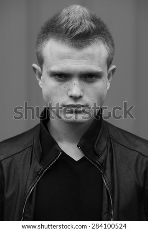 Stylish bully concept. Portrait of brutal young man with short hair wearing black jacket and posing over urban background. Hipster style. Close up. Black and white outdoor shot