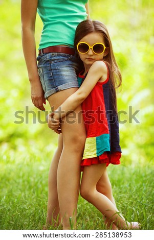 Happy weekend concept. Portrait of fashionable gorgeous baby girl in trendy sunglasses hugging her mother. Family walking in the park. Stylish casual clothes. Sunny summer day. Outdoor shot
