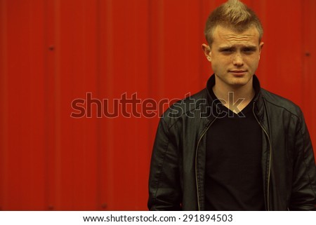 Stylish bully concept. Portrait of brutal young man with short blond hair wearing black jacket and posing over red urban background. Hipster style. Close up. Copy-space. Outdoor shot