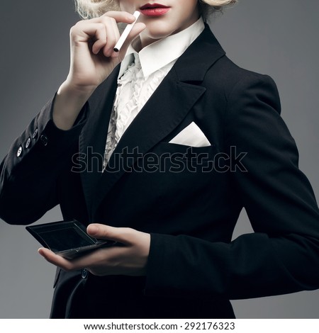 Femme fatale concept. Marlene Dietrich style. Close up retro portrait of rich young woman smiling wearing expensive luxurious tuxedo and smoking cigarette over gray background. Studio shot