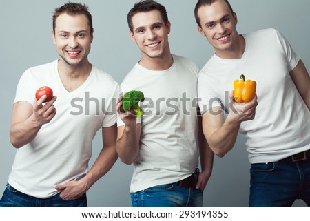 Raw, living food, veggie concept. Portrait of three happy young men wearing white t-shirts, blue jeans, holding vegetables in hands over gray background. Casual clothing. Muscular bodies. Studio shot