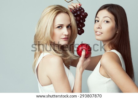 Raw, living food, veggie concept. Portrait of two happy young women wearing white sleeveless shirts, holding fruits over gray background. Casual clothing. Perfect skin, natural make-up. Copy-space