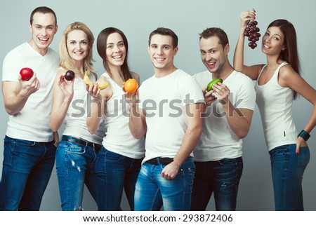 Raw, living food, veggie concept. Group portrait of healthy boys, girls in white t-shirts, sleeveless shirts and blue jeans standing with fruits, posing over gray background. Urban style. Studio shot