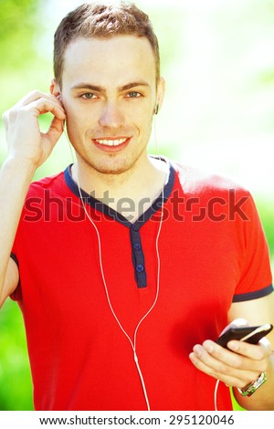 Gadget user concept. Portrait of smiling guy in red casual t-shirt using earphones in park. White shiny smile. Bristle on face with healthy skin. Urban style. Close up. Outdoor shot